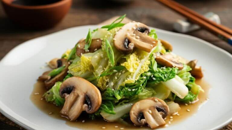 A vibrant plate of Chinese cabbage stir-fry with sliced mushrooms, glistening with a light soy-based sauce. The dish is served on a white plate, garnished with fresh green onions and sesame seeds, surrounded by chopsticks and a small bowl of soy sauce on a wooden table.