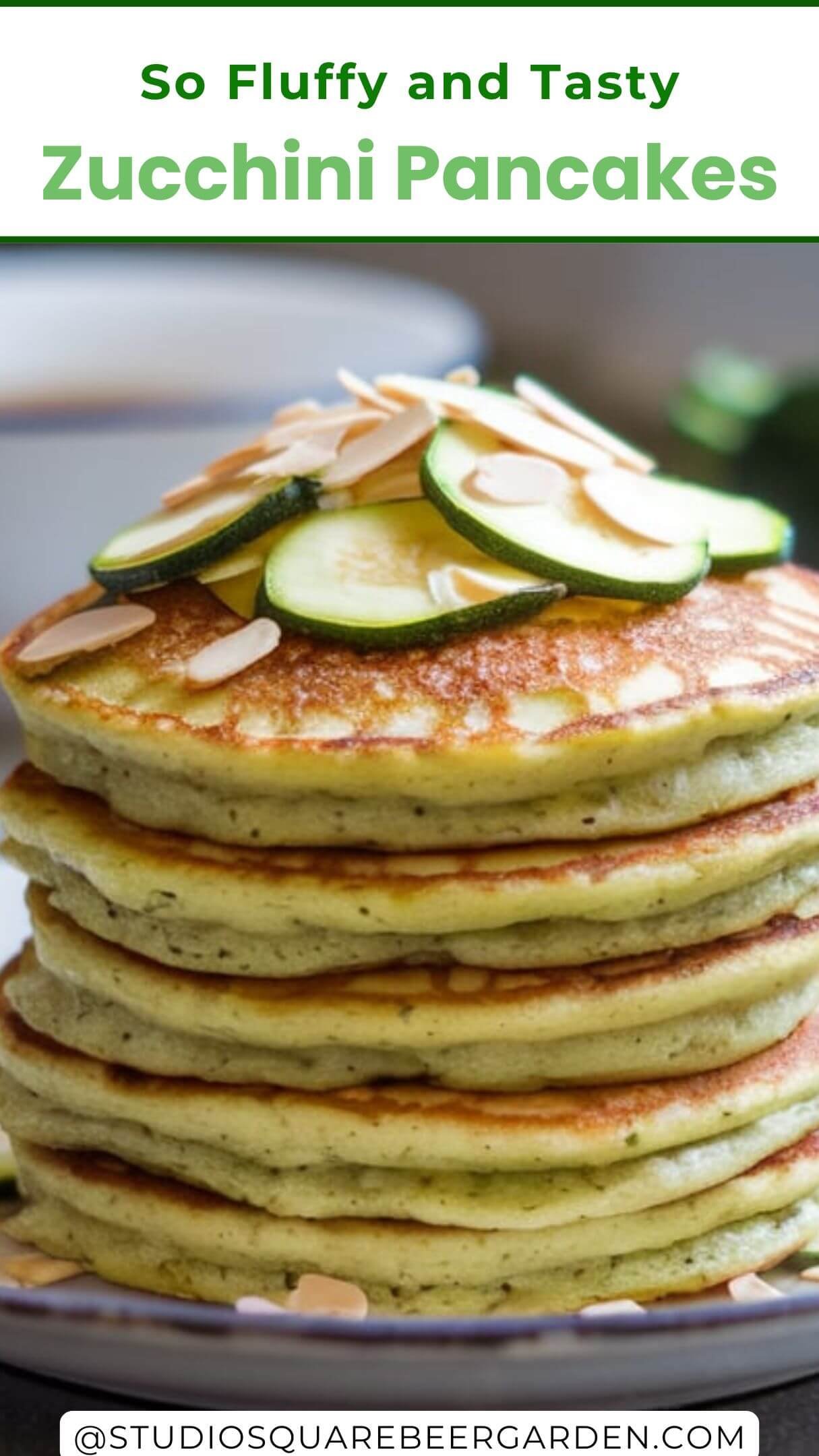 A stack of Zucchini pancakes topped with thinly sliced zucchini and almond slices. The pancakes are golden brown and appear to be freshly cooked. They are placed on a white plate, which is set on a gray countertop. In the background, there's a white bowl, possibly containing syrup or another topping, and a whole zucchini. The setting is kitchen, with a natural light