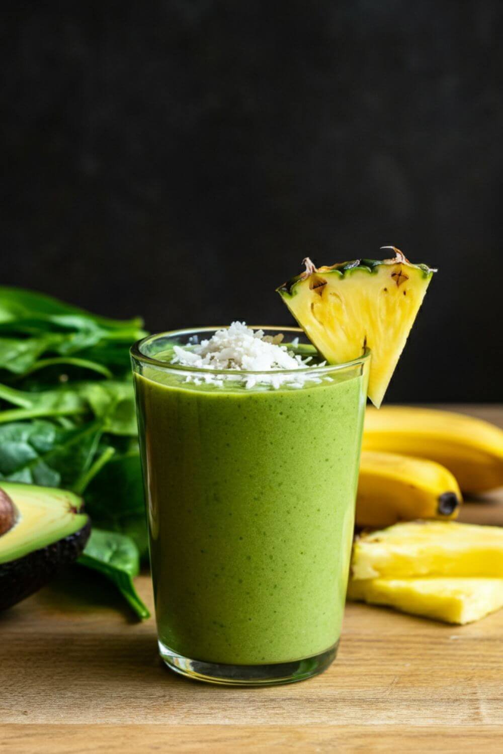 A vibrant green smoothie served in a clear glass, garnished with a small pineapple wedge and a sprinkle of shredded coconut. The smoothie is creamy and visually refreshing, with a tropical vibe. Place the glass on a wooden table surrounded by fresh spinach leaves, slices of pineapple, half a banana, an avocado, and a small jar of coconut milk for a natural and wholesome aesthetic. Bright, natural lighting enhances the freshness of the ingredients.