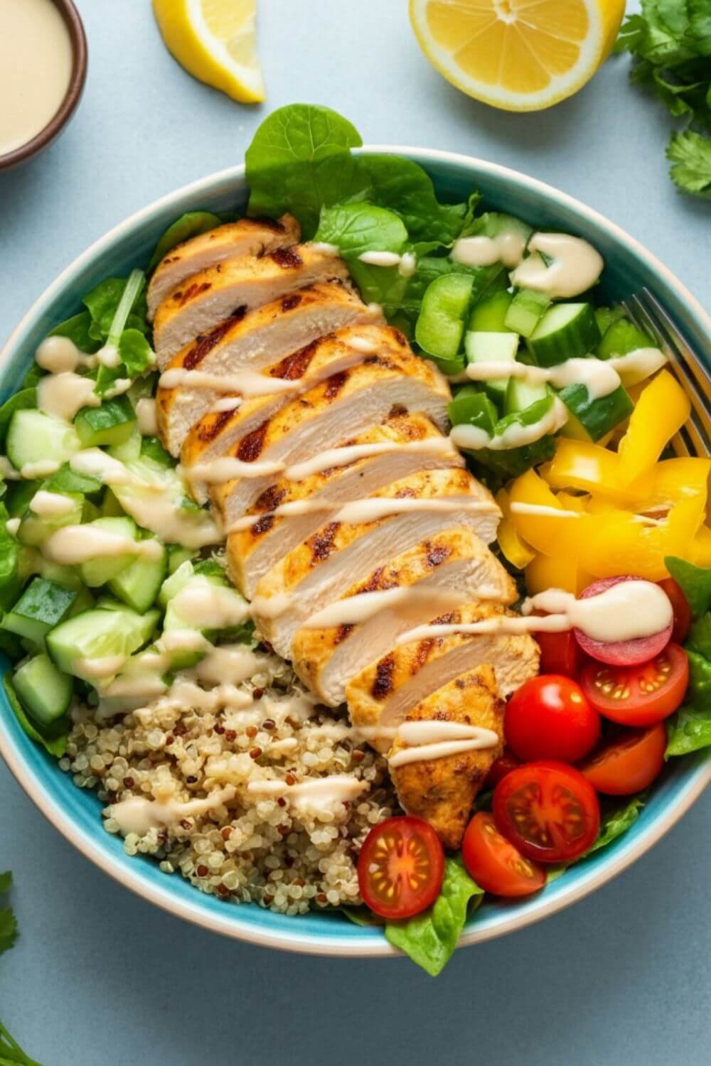 A vibrant salad bowl featuring sliced grilled chicken over a bed of quinoa and colorful fresh veggies, garnished with a creamy lemon-tahini dressing drizzled on top. The background is light and airy, with fresh ingredients scattered around for a wholesome, inviting vibe.