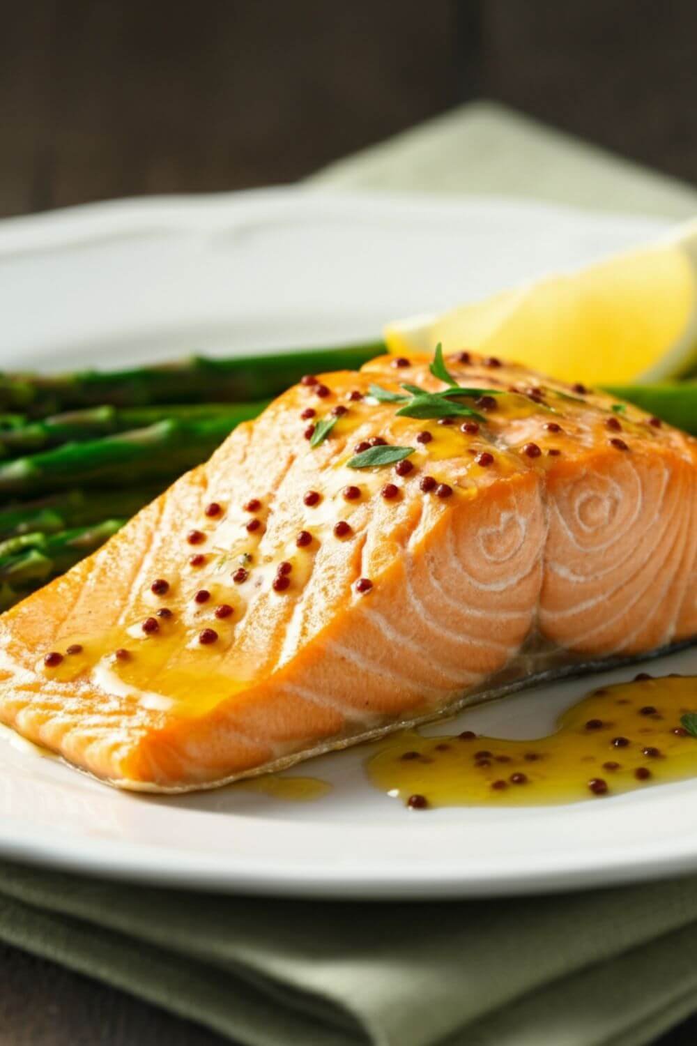 A golden-brown baked salmon fillet glistening with honey mustard glaze, garnished with a lemon wedge and paired with vibrant green asparagus on a white plate. Fresh herbs and a light drizzle of sauce decorate the scene.