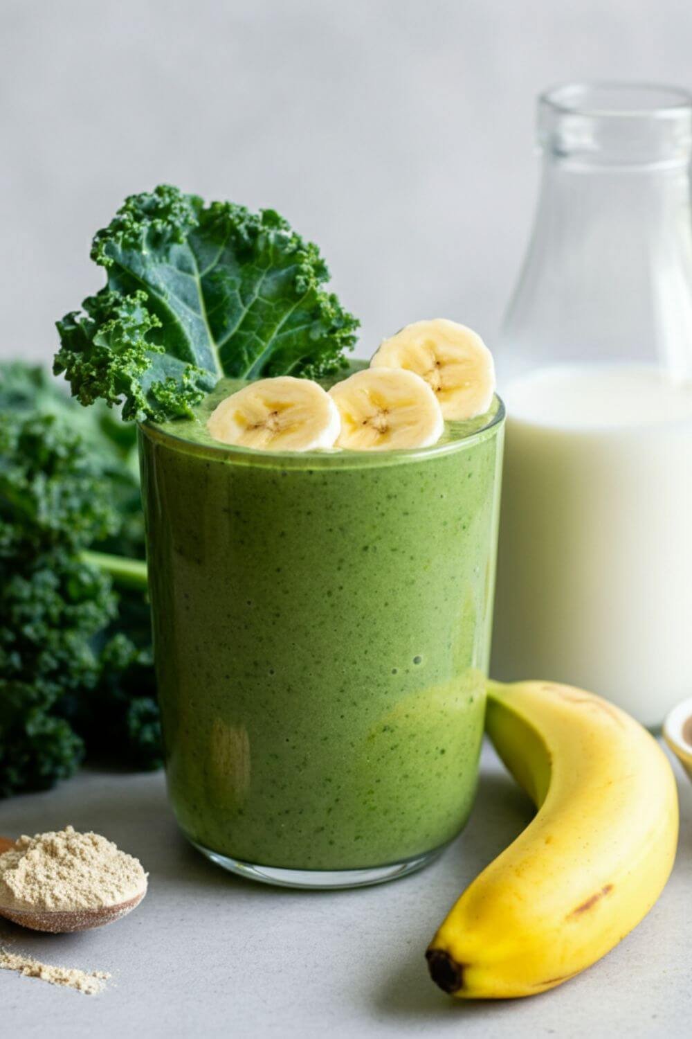 A vibrant green smoothie in a clear glass, garnished with a small kale leaf and banana slices. Surround the glass with the ingredients: fresh kale leaves, half a banana, a scoop of protein powder in a small dish, a spoonful of almond butter, and a glass of almond milk. Bright, natural lighting with a clean, minimal background to emphasize freshness and healthiness.