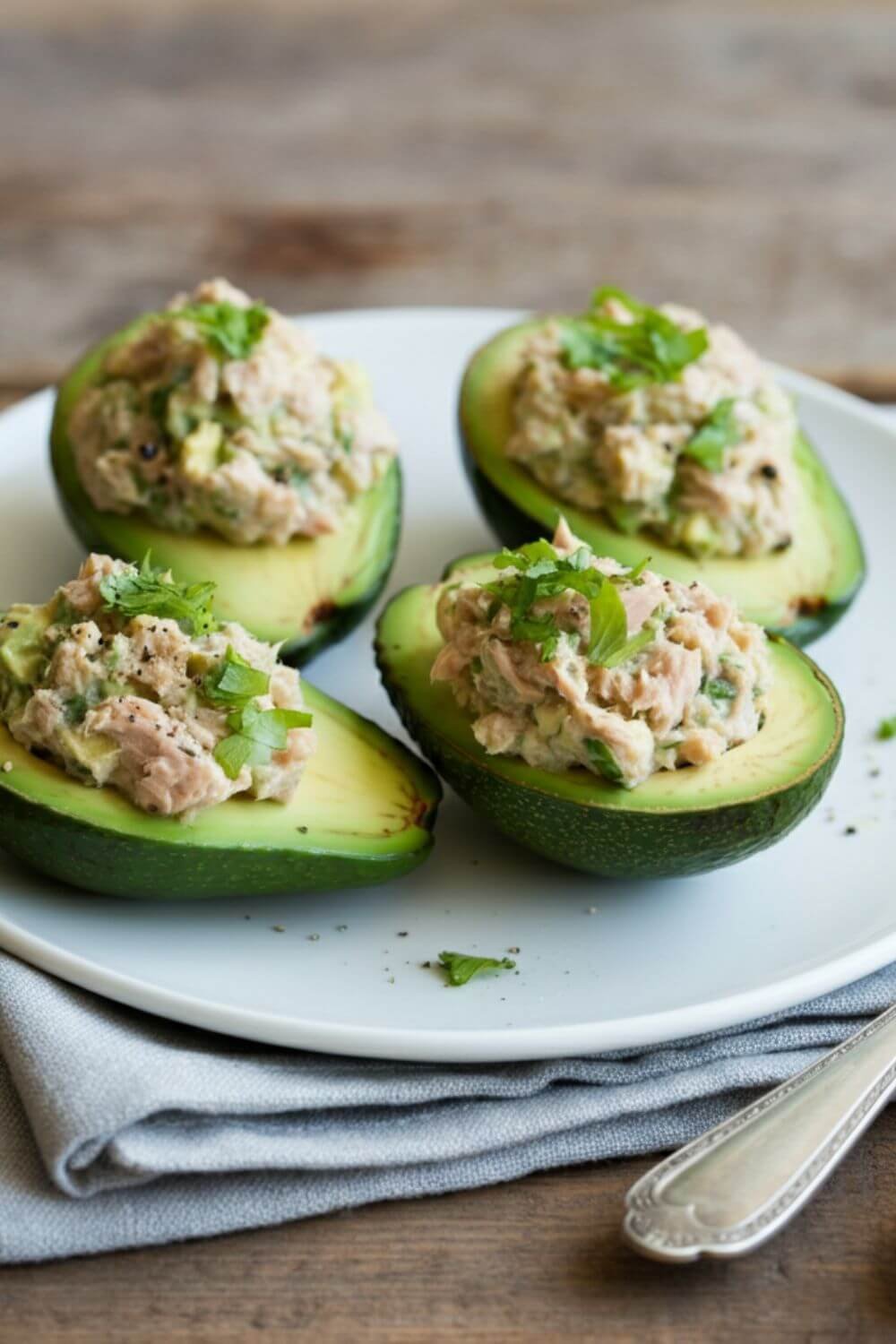 A vibrant image of 4 avocado halves on a white plate, filled with a creamy tuna mixture and garnished with freshly cracked pepper and a sprinkle of herbs. The background features a wooden table with a fork and napkin nearby.