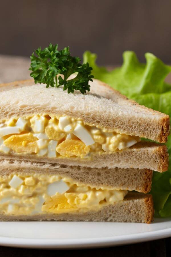 A close-up of a simple egg salad sandwich on whole-grain bread, cut diagonally with creamy filling spilling slightly out the edges. Garnish with a sprig of parsley and a side of fresh lettuce leaves. Placed in the white dish.