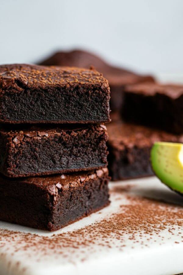 A close-up of rich, fudgy brownies with a glossy top, arranged on a white board. Garnish with a sprinkle of cocoa powder and a slice of fresh avocado on the side for a hint of the secret ingredient.