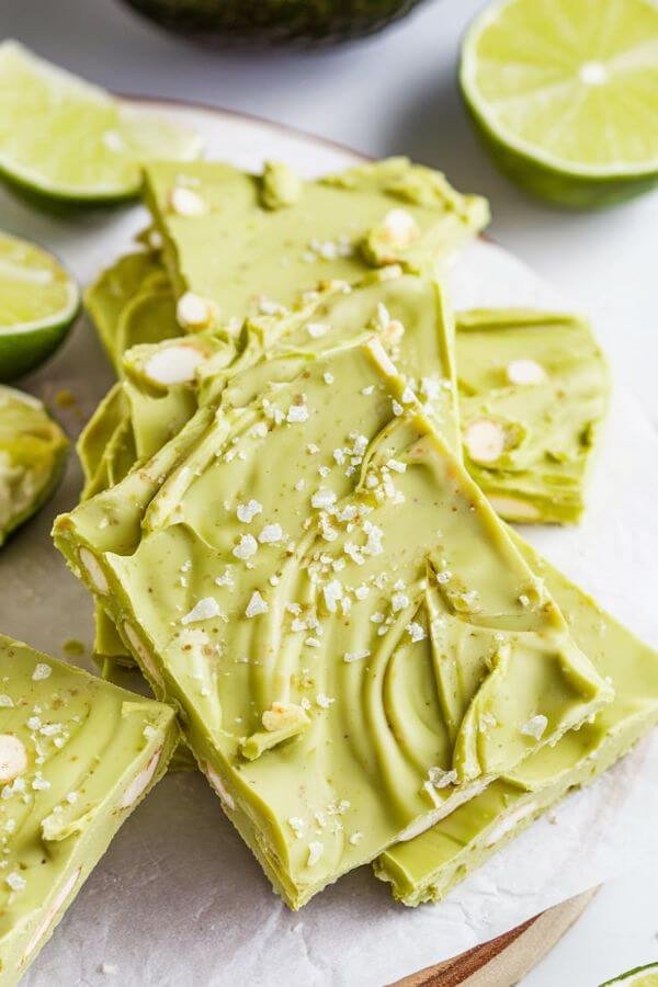 A close-up of vibrant green bark with a creamy texture, speckled with bits of white chocolate and a dash of salt flakes, served on a white board with sliced limes and an avocado in the background.