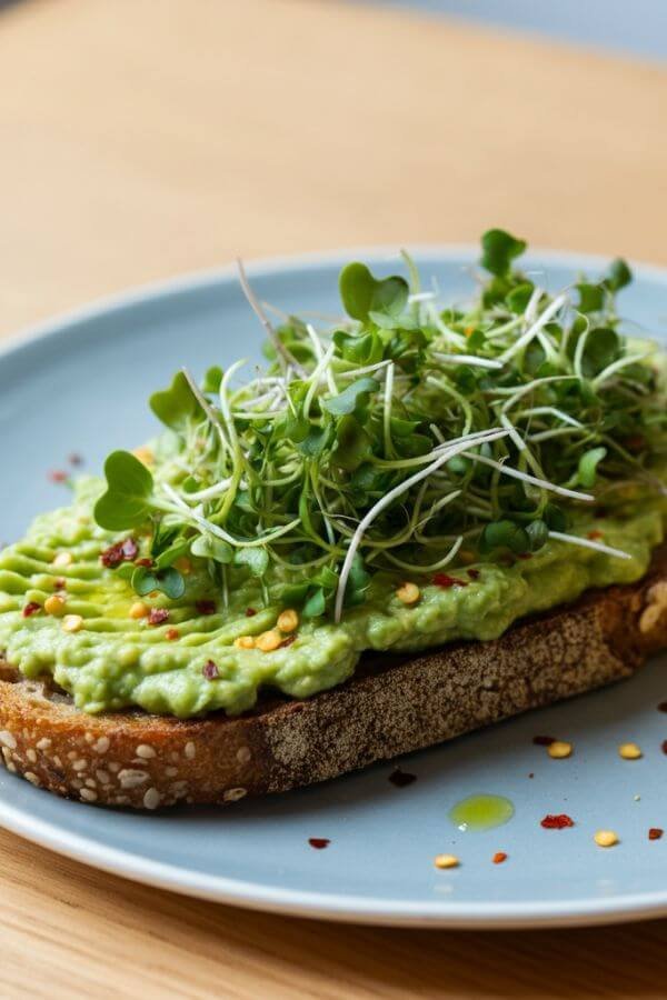 2 beautifully slice of golden sourdough toast covered in creamy avocado spread, topped with fresh vibrant microgreens and a dash of chili flakes, set on a white dish with a lemon wedge on the side.