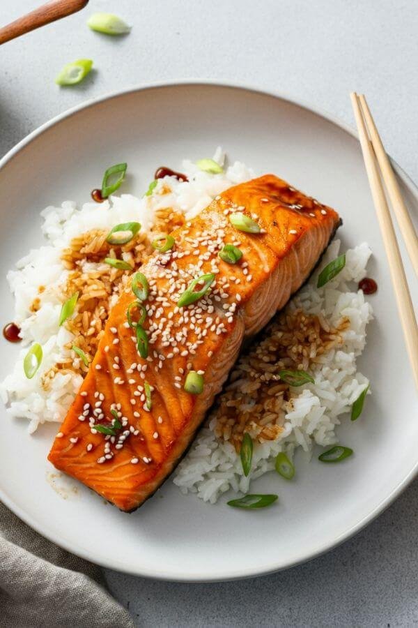 A beautifully plated dish of golden, caramelized teriyaki salmon fillets glistening with sauce, served over a bed of fluffy white rice, garnished with sesame seeds and green onions. A small ramekin of extra teriyaki glaze and chopsticks lie nearby on a white table.