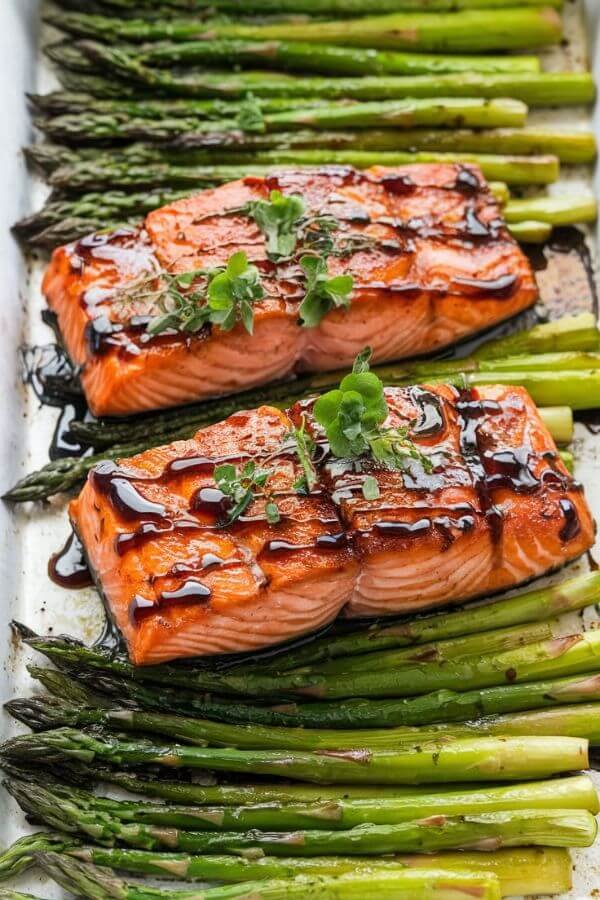 A beautifully white sheet pan of balsamic-glazed salmon resting on a bed of roasted asparagus, garnished with a drizzle of glaze and fresh herbs. The salmon has a caramelized, glossy finish, while the asparagus is bright green with a slight char.