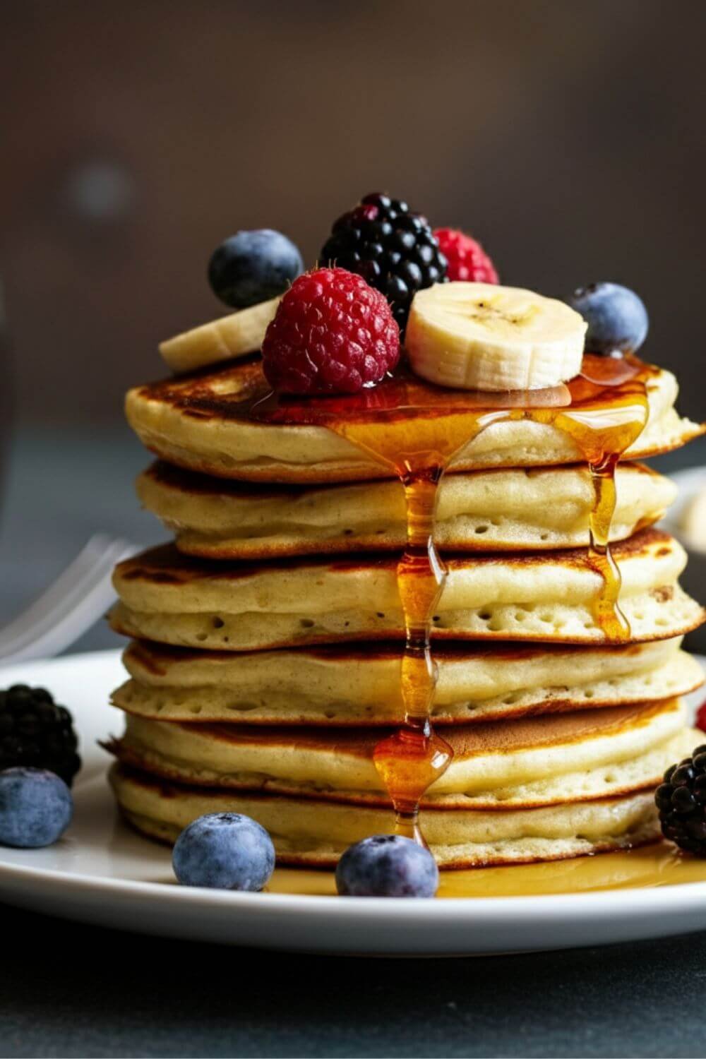 A stack of golden, fluffy banana pancakes on a white plate, topped with fresh fruit (like berries or banana slices) and a drizzle of maple syrup, with a light background.