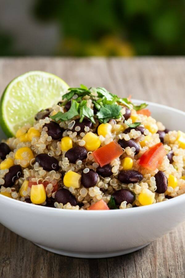 A white bowl filled with fluffy quinoa, black beans, sweet corn kernels, and fresh diced tomatoes, topped with a sprinkle of chopped cilantro. A lime wedge sits on the side of the bowl, highlighting the freshness of the dish. The setting is bright and natural, with a rustic wooden table and a hint of greenery in the background.