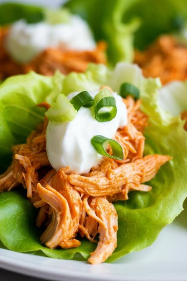 A close-up of 4 fresh green lettuce wraps filled with bright shredded buffalo chicken, topped with a dollop of creamy Greek yogurt and garnished with chopped celery and green onions. Serve on a clean white plate for a vibrant, healthy presentation.