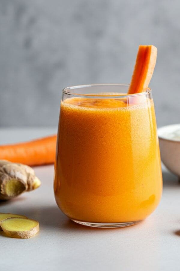 A bright orange smoothie in a glass, garnished with a carrot stick or a small piece of fresh ginger, placed on a white table with fresh carrots, ginger, and a bowl of yogurt nearby.