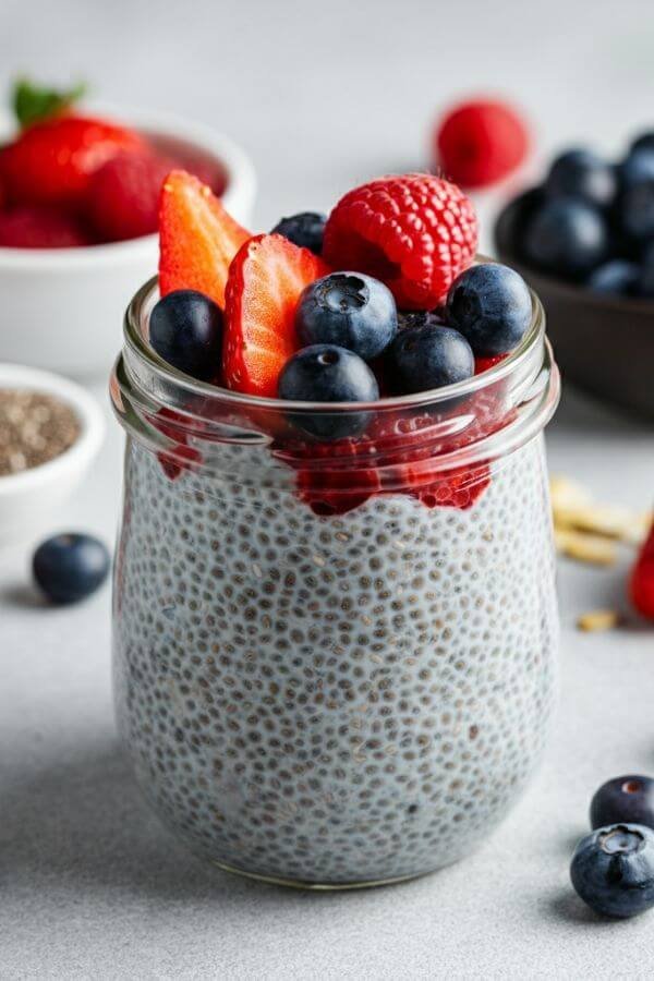 A vibrant, close-up shot of a glass jar filled with creamy chia seed pudding, topped with an assortment of fresh berries like blueberries, raspberries, and sliced strawberries. The pudding has a smooth, thick texture with visible chia seeds throughout. The jar is placed on a white surface, surrounded by scattered fresh berries, a small bowl of chia seeds, and a drizzle of honey or maple syrup in the background. Soft natural lighting enhances the freshness and colors of the ingredients.