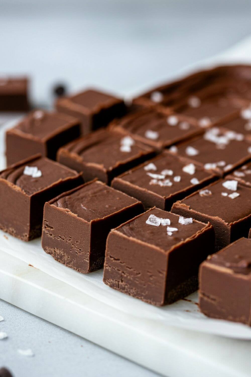 A square pan of silky chocolate fudge, cut into small squares, with a light sprinkle of sea salt on top. Displayed on a white board with a few scattered chocolate chips around. the background is light