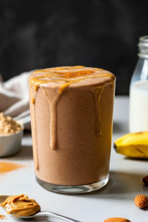 A glass of creamy chocolate peanut butter smoothie placed on a white countertop. The glass is garnished with a slight drizzle of peanut butter and a sprinkle of cinnamon. Surround it with the key ingredients: a scoop of protein powder, a banana slice, a spoonful of peanut butter, and a small jar of almond milk. Keep the lighting natural and warm for a cozy, inviting look.