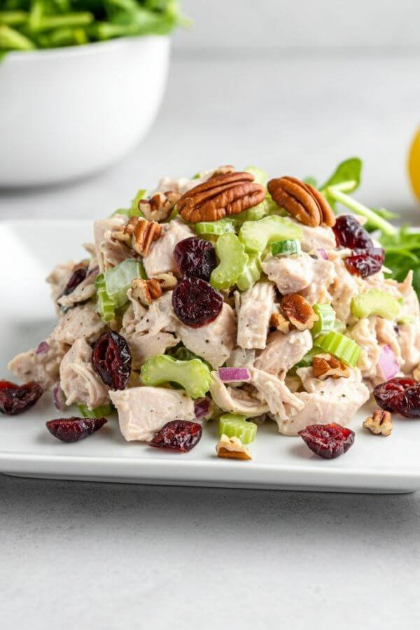 A vibrant, fresh chicken salad served on a white dish, with pieces of dried cranberries, chopped pecans, and diced celery visible. The dish is styled on a white table with a side of fresh greens and a lemon wedge in the background.