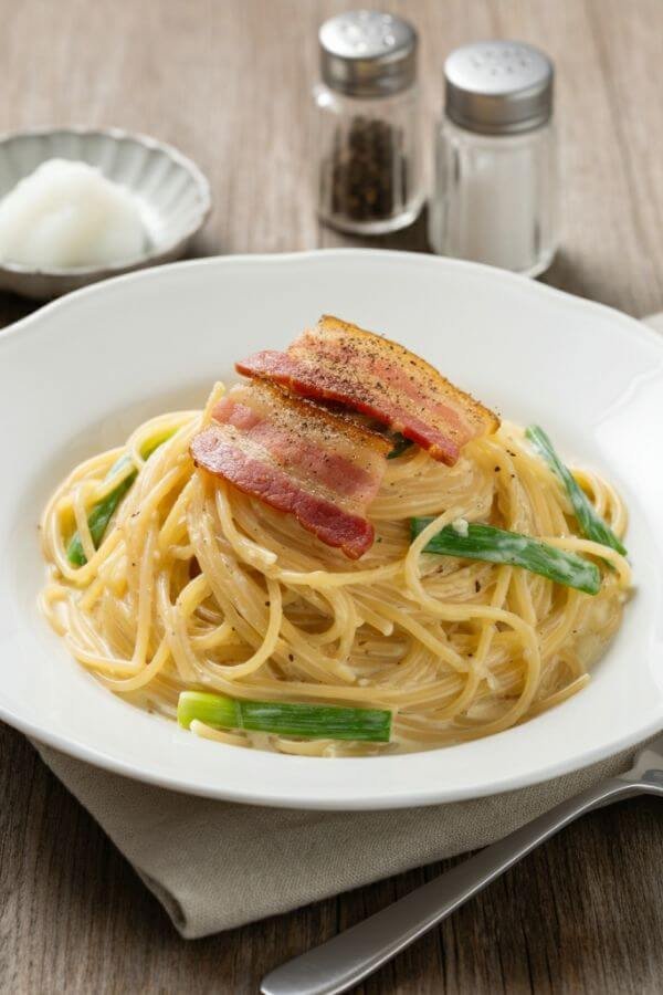 A white plate of creamy spaghetti topped with golden crispy pancetta, freshly cracked black pepper, and vibrant green scallions, arranged neatly on a rustic table with a small dish of white miso and a fork on the side. the background is light and airy, giving a cozy yet modern feel. 