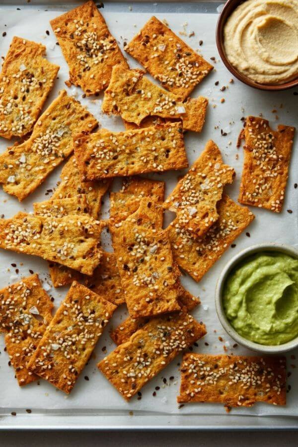 A tray of golden-brown sourdough discard crackers on parchment paper, sprinkled with sea salt and sesame seeds, with small bowls of hummus and guacamole on the side.