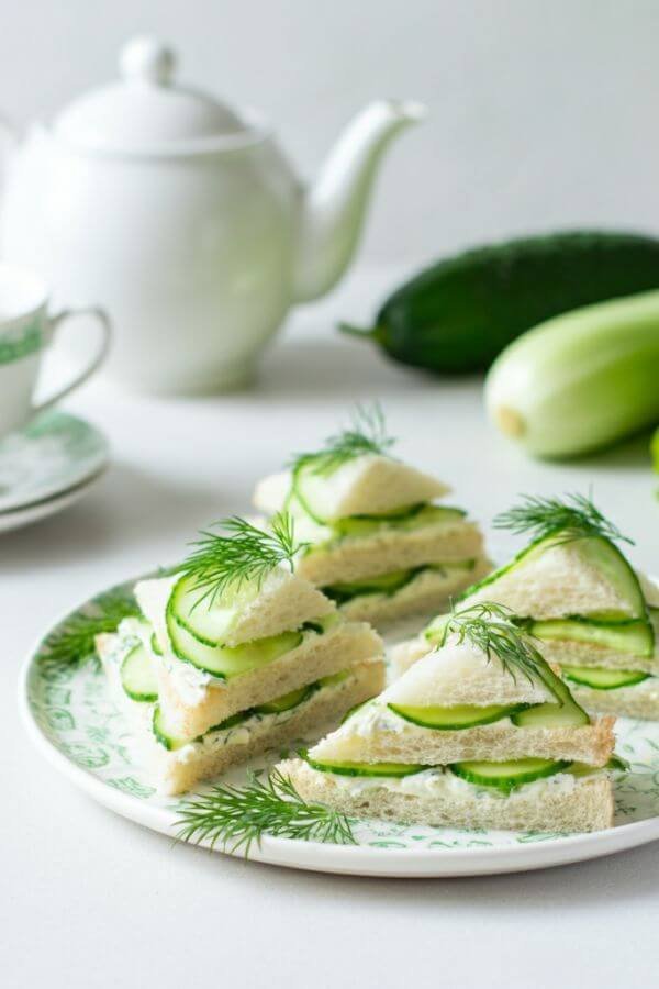 A plate of small, triangle-cut cucumber sandwiches arranged neatly. The sandwiches should show layers of green cucumber and creamy cheese, garnished with fresh dill sprigs. Set against a bright, clean background with a tea set or fresh veggies nearby for an inviting look.