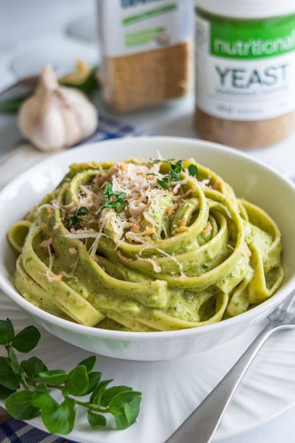 A vibrant bowl of edamame fettuccine Alfredo topped with a sprinkle of Parmesan and fresh herbs, set against a WHITE with soft, natural lighting. The creamy sauce glistens, and steam rises gently, highlighting its warmth. Include a fork resting on the side and some fresh garlic or nutritional yeast as props in the background.