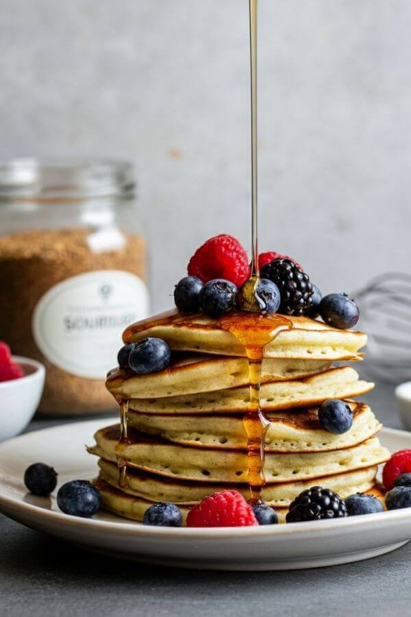 A stack of golden-brown pancakes topped with fresh berries, a pat of melting butter, and a drizzle of maple syrup. Show a rustic kitchen setting with a small jar of sourdough discard and a whisk in the background.