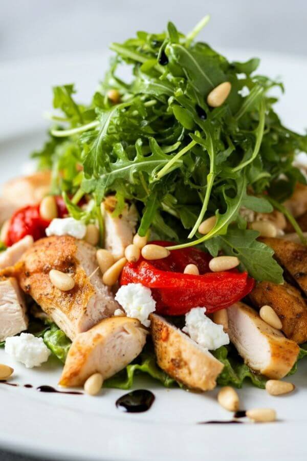 A gourmet chicken salad on a white dish, topped with fresh arugula or microgreens. The salad should have visible chunks of roasted chicken, crumbled goat cheese, vibrant red peppers, and golden pine nuts, with a slight drizzle of truffle oil glistening on top. Add soft, natural lighting for a fresh and appetizing look. the chicken is chopped