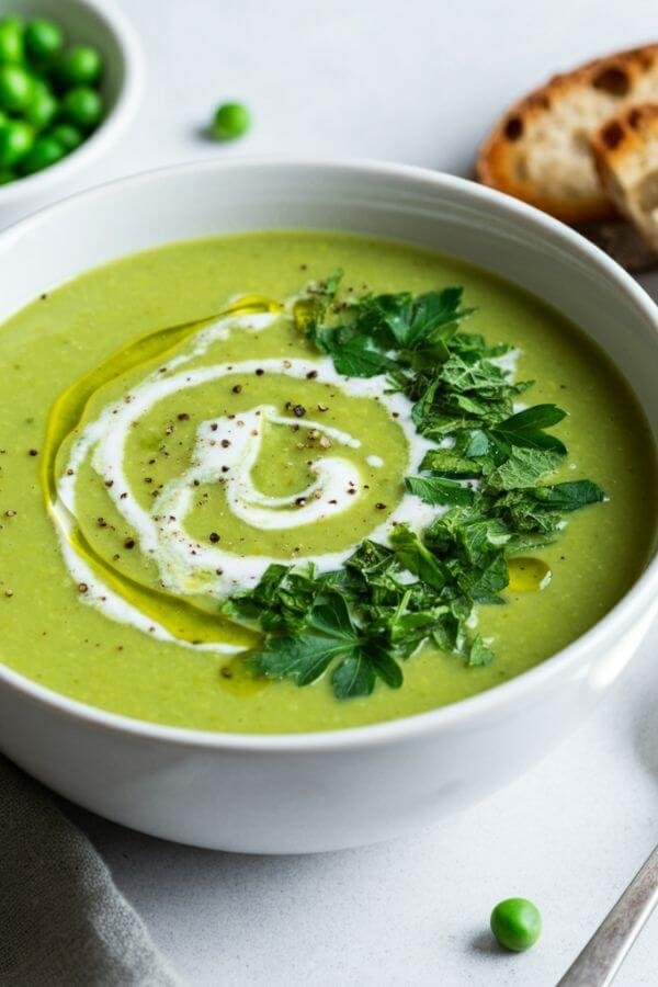 A vibrant bowl of creamy green pea soup served in a white bowl, garnished with a swirl of coconut milk, fresh herbs like parsley or mint, and a sprinkle of cracked black pepper. Place the bowl on a white table with a spoon and slices of crusty bread on the side.