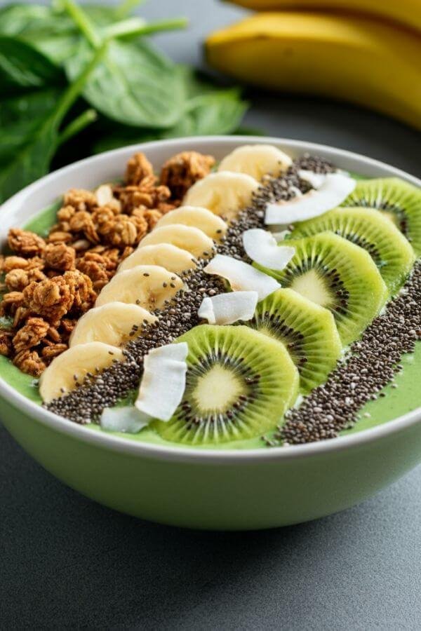 A vibrant green smoothie bowl topped with granola, sliced kiwi, chia seeds, and coconut flakes, styled on a table with fresh spinach and bananas in the background. the background is light