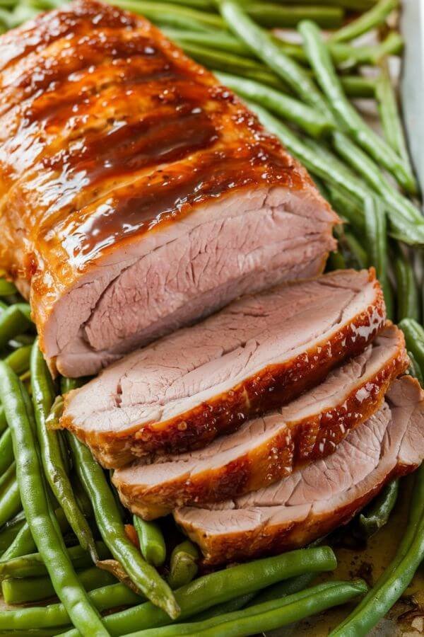 A beautifully roasted pork tenderloin with a glossy hoisin glaze, sliced to reveal the juicy interior, resting on a bed of vibrant green beans. The golden-brown caramelization on the pork contrasts with the bright, fresh colors of the green beans, all presented on a white sheet pan.