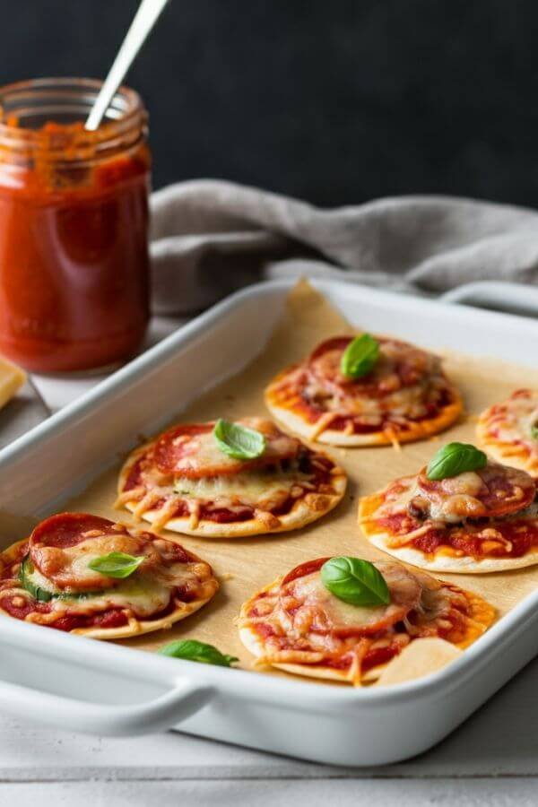 A white baking tray with mini pizzas made from tortillas, topped with melted cheese, colorful veggies, and pepperoni. The pizzas are garnished with fresh basil and placed on a white table next to a jar of marinara sauce and a block of cheese.