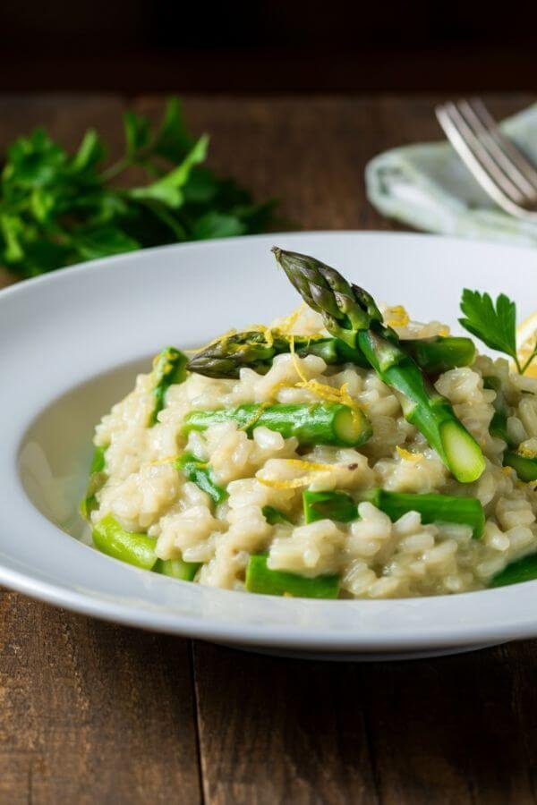 A vibrant plate of creamy risotto with bright green asparagus tips and a sprinkle of lemon zest on top. The dish is served in a white bowl with a wedge of lemon and a sprig of parsley on the side for garnish.