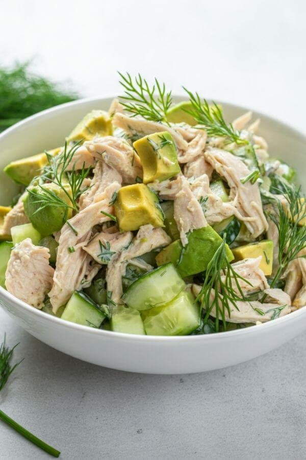 A vibrant, fresh chicken salad served in a white bowl. The salad features chunks of chicken, diced cucumber, creamy avocado, and a sprinkle of fresh dill, all lightly coated in a creamy dressing. The background is minimalistic, with a white surface and a few sprigs of dill lying nearby. The focus is on the fresh, colorful ingredients.