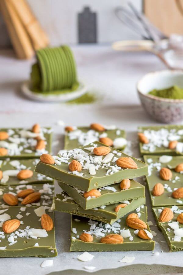 A thin layer of glossy green matcha chocolate bark sprinkled with white coconut flakes and golden-brown almond chunks, set on a parchment-lined tray. The light background features a cozy kitchen with matcha powder and white chocolate in small bowls.