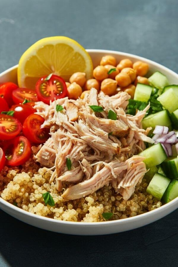 A vibrant Mediterranean bowl with shredded chicken on a bed of quinoa, surrounded by colorful cherry tomatoes, chopped cucumber, red onion, and chickpeas. Garnish with a slice of lemon and a sprinkle of fresh herbs for a fresh, wholesome look.