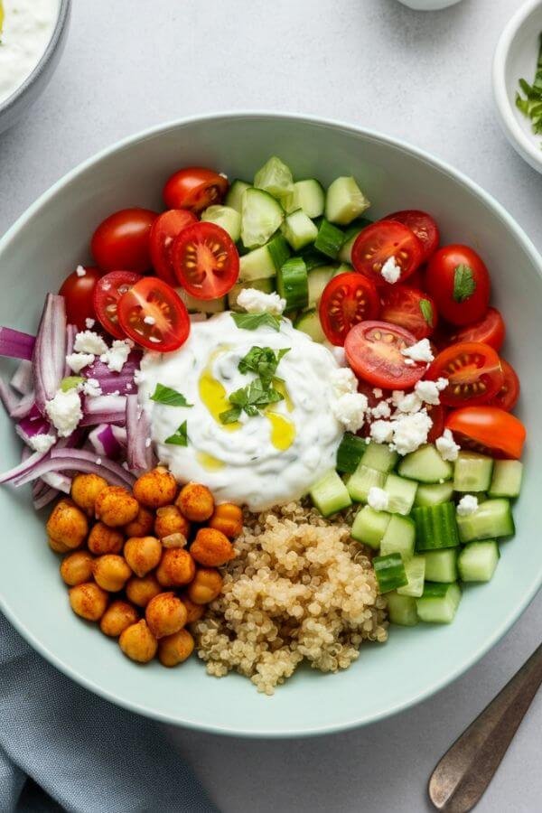 A vibrant Mediterranean rice bowl with a base of fluffy quinoa, surrounded by spiced chickpeas, bright cherry tomatoes, cucumbers, and red onions, topped with creamy tzatziki, a drizzle of olive oil, and a sprinkle of feta cheese. The bowl is placed on a white table with a small dish of tzatziki and fresh herbs on the side.