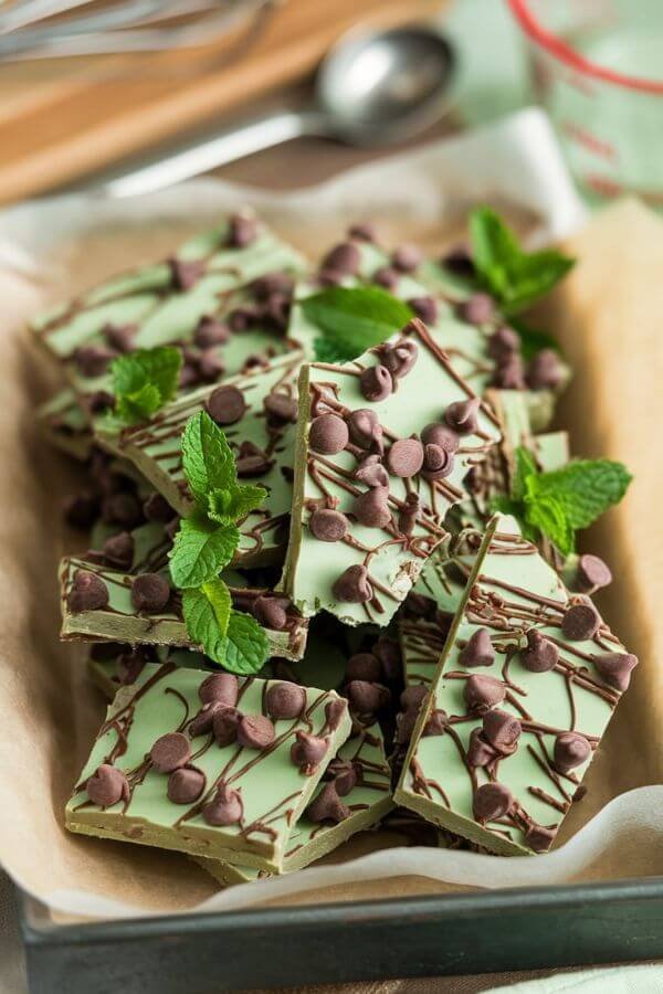 A bright and vibrant tray of frozen green bark broken into rustic pieces. The bark has flecks of fresh mint leaves and dark chocolate chips, laid on parchment paper with a soft-focus background of kitchen elements. The colors are fresh and inviting, highlighting the creamy green texture and rich chocolate.