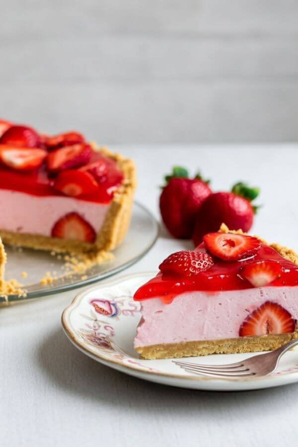 A vibrant no-bake strawberry pie sitting on a white table. The pie features a golden graham cracker crust, a creamy white filling, and fresh, glossy strawberries piled on top with a light glaze. A few scattered strawberries and a slice of pie on a plate with a fork complete the scene.