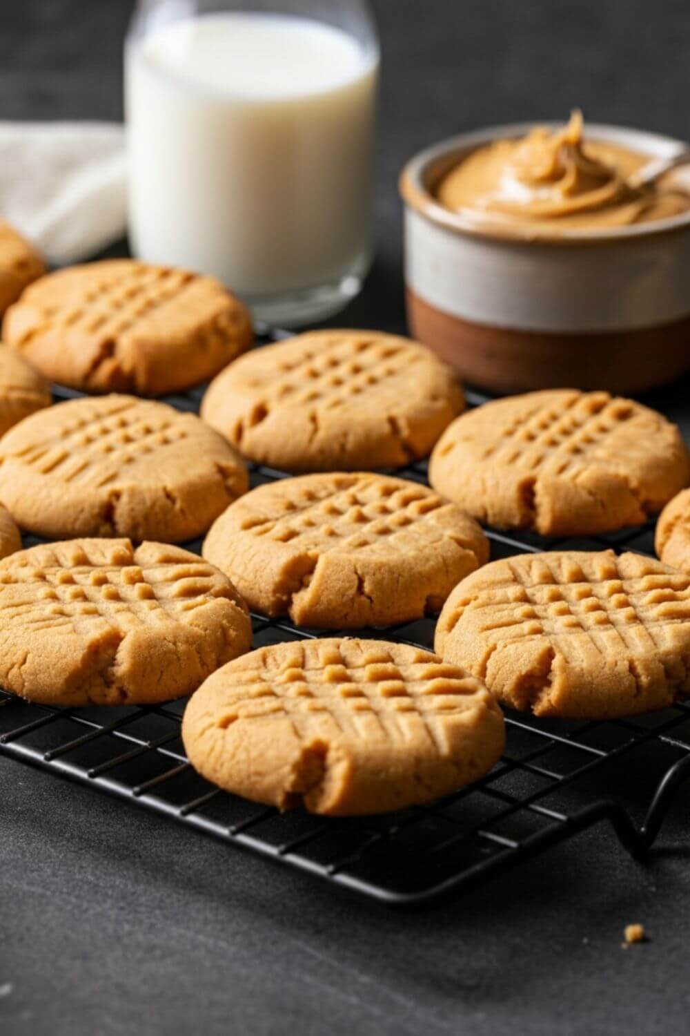 A batch of golden-brown peanut butter cookies arranged on a cooling rack, with a crisscross fork pattern on top. Include a small bowl of peanut butter and a glass of milk in the light background for a cozy touch.