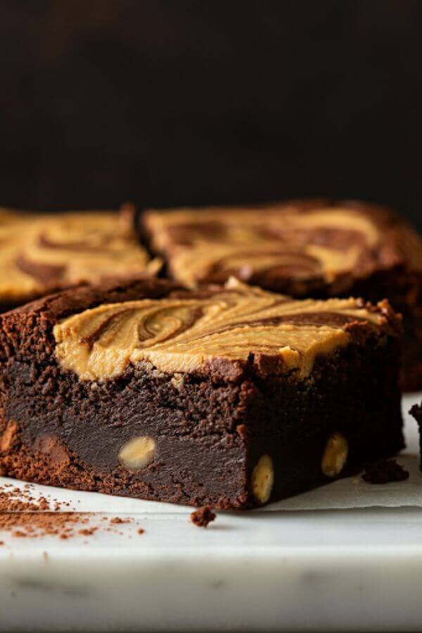 A close-up of gooey, fudgy brownies with a golden peanut butter swirl on top. They're cut into squares, placed on a white board, with a small jar of peanut butter and a cocoa-dusted spoon in the background.