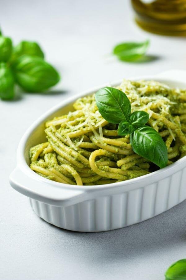 A bowl of vibrant green pesto pasta served in a white ceramic dish, garnished with a fresh basil sprig and a sprinkle of grated Parmesan. The background features a white table with scattered basil leaves, a wedge of Parmesan, and a small jar of olive oil for a cozy, rustic vibe.