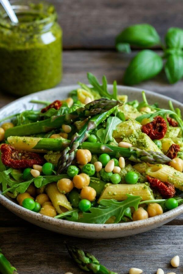 A bowl of vibrant green pasta salad featuring chickpea pasta coated in pesto, mixed with asparagus, peas, arugula, sun-dried tomatoes, and sprinkled with pine nuts. Place it on a wooden table with fresh basil leaves and a jar of pesto in the background.