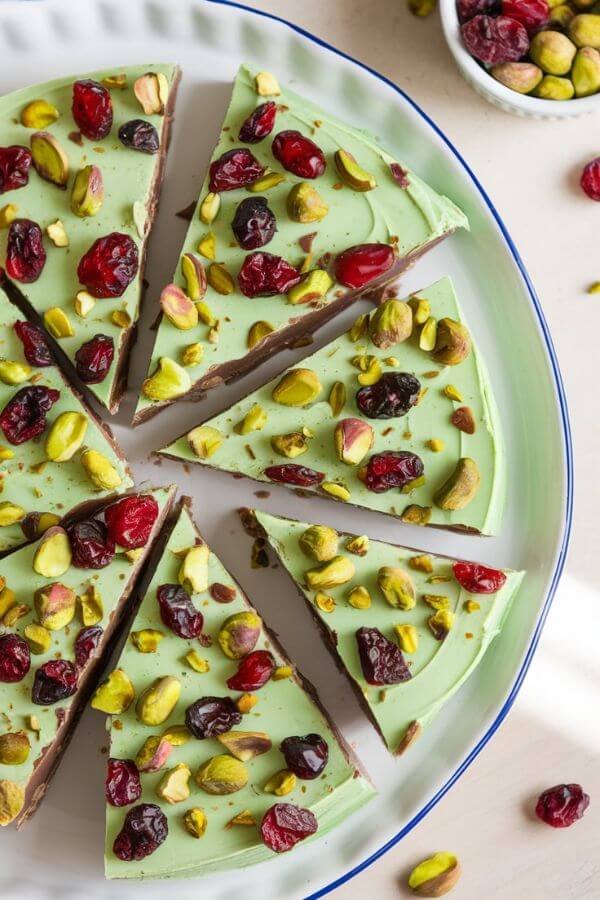 A flat-lay shot of Pistachio Cranberry Green Bark on a white dish. The bark features a vibrant green yogurt base, topped with scattered red dried cranberries and chopped green pistachios, creating a festive and colorful look. Pieces of the bark are broken apart artistically, with a small bowl of pistachios and cranberries placed nearby for added texture. Soft natural lighting highlights the creamy and crunchy textures, evoking a fresh and wholesome feel.