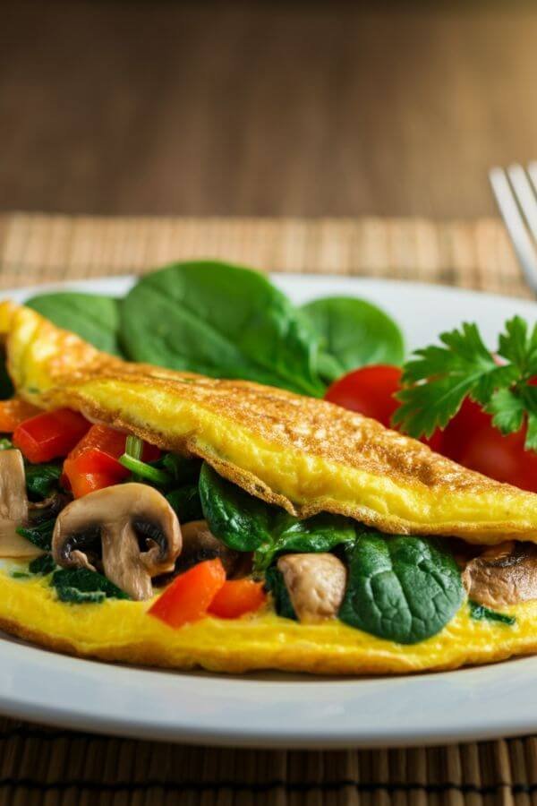 A vibrant veggie omelette on a white plate, garnished with fresh parsley and paired with a side of cherry tomatoes. The omelette should showcase colorful veggies like spinach, mushrooms, and bell peppers peeking through the golden egg base.