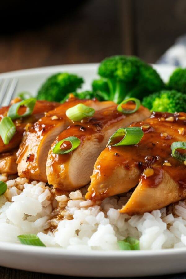 A close white plate of tender, glazed chicken breasts covered in a rich honey garlic sauce, served over a bed of fluffy white rice with a sprinkle of green onions on top. Add clear steamed broccoli for a complete, colorful meal.