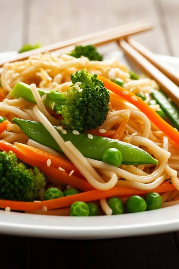 A close lok of stir-fried noodles in a white dish, with colorful veggies like carrots, broccoli, and peas. Garnished with sesame seeds, served with chopsticks on the side.