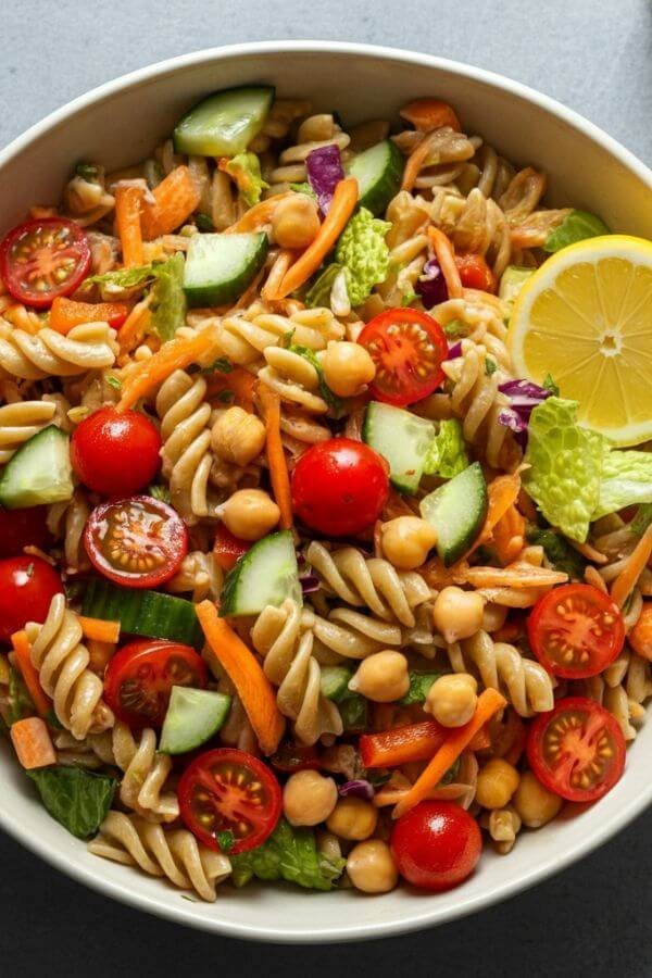 A vibrant overhead shot of a colorful pasta salad in a large bowl. The salad should feature bright, fresh veggies like cherry tomatoes, cucumbers, shredded carrots, and bell peppers mixed with whole-grain pasta. Include a drizzle of olive oil and a lemon wedge on the side for a fresh, natural look. Optional protein like chickpeas or grilled chicken can be scattered on top.