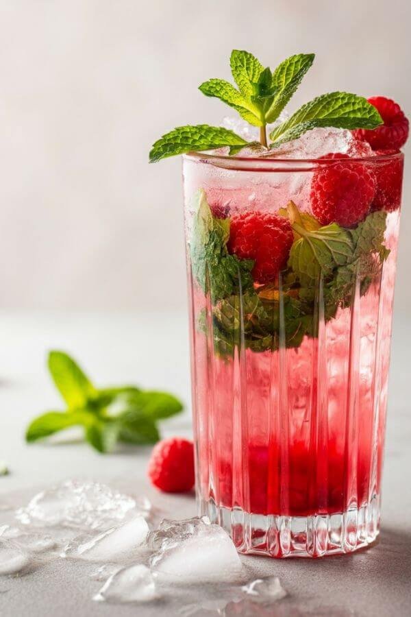 A vibrant Raspberry Mojito in a tall glass, filled with crushed ice, topped with mint and fresh raspberries. The glass is placed on a light background with scattered mint leaves and raspberries around for a fresh, festive vibe.