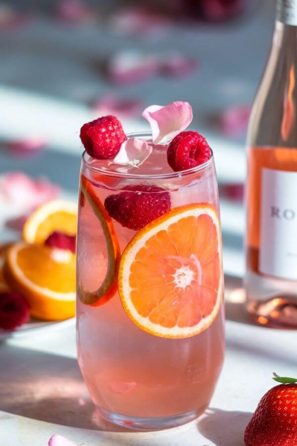 A tall glass filled with pale pink sangria, garnished with vibrant slices of orange and strawberries. Fresh raspberries float on top, alongside a few delicate edible rose petals. The scene is set on a light white table with soft natural lighting, surrounded by scattered petals and a bottle of rosé wine in the background.