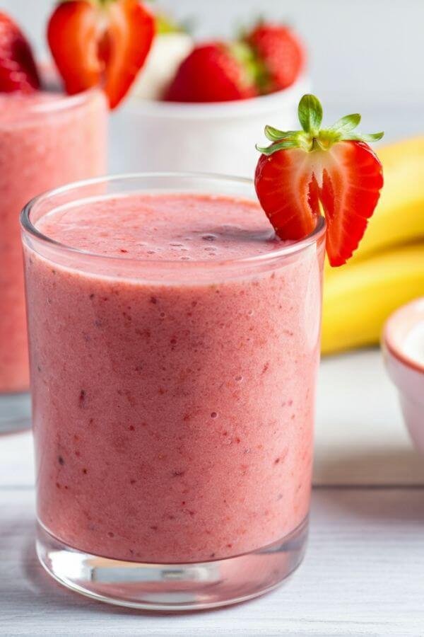 A bright, refreshing strawberry smoothie in a clear glass, topped with a strawberry slice on the rim. The light background includes fresh strawberries, a banana, and a small bowl of Greek yogurt.